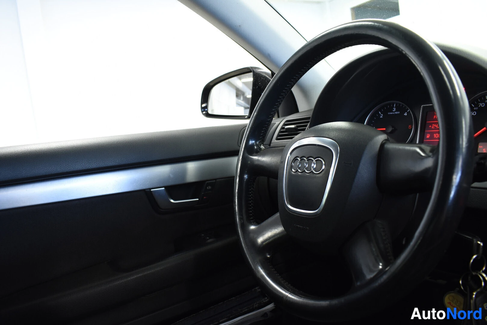 Ohjaamo: Musta nahkainen ohjauspyörä, jossa on Audi-logo keskellä, näkyy vasemmalta puolelta. Mittaristo näkyy osittain taustalla. Vasen oven sisäpuoli on kuvassa, jossa