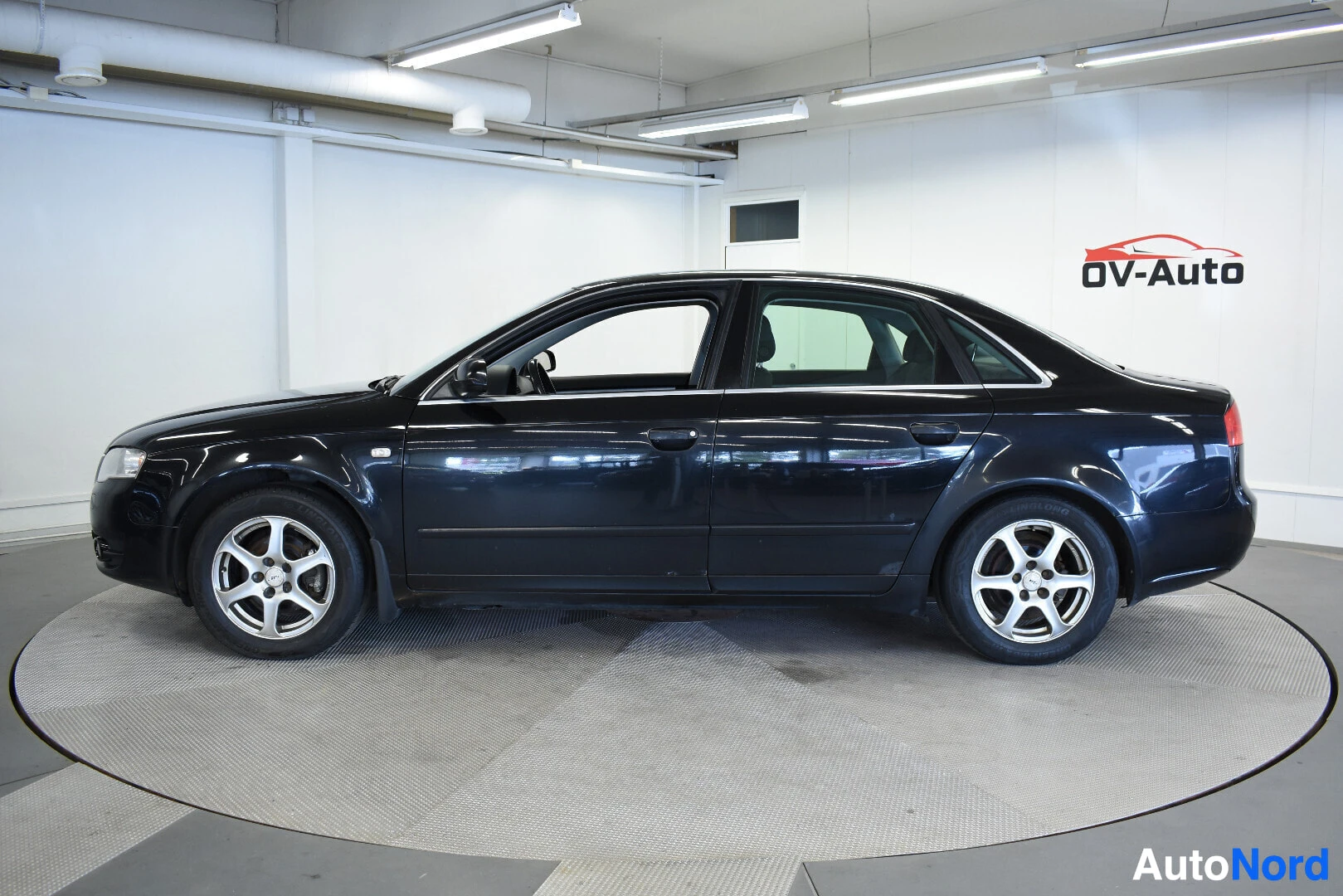 Sivulta: Musta Audi A4 -henkilöauto vasemmalta sivulta. Autossa on viisipuolaiset kevytmetallivanteet. Taustalla valkoinen seinä ja OV-Auton logo.