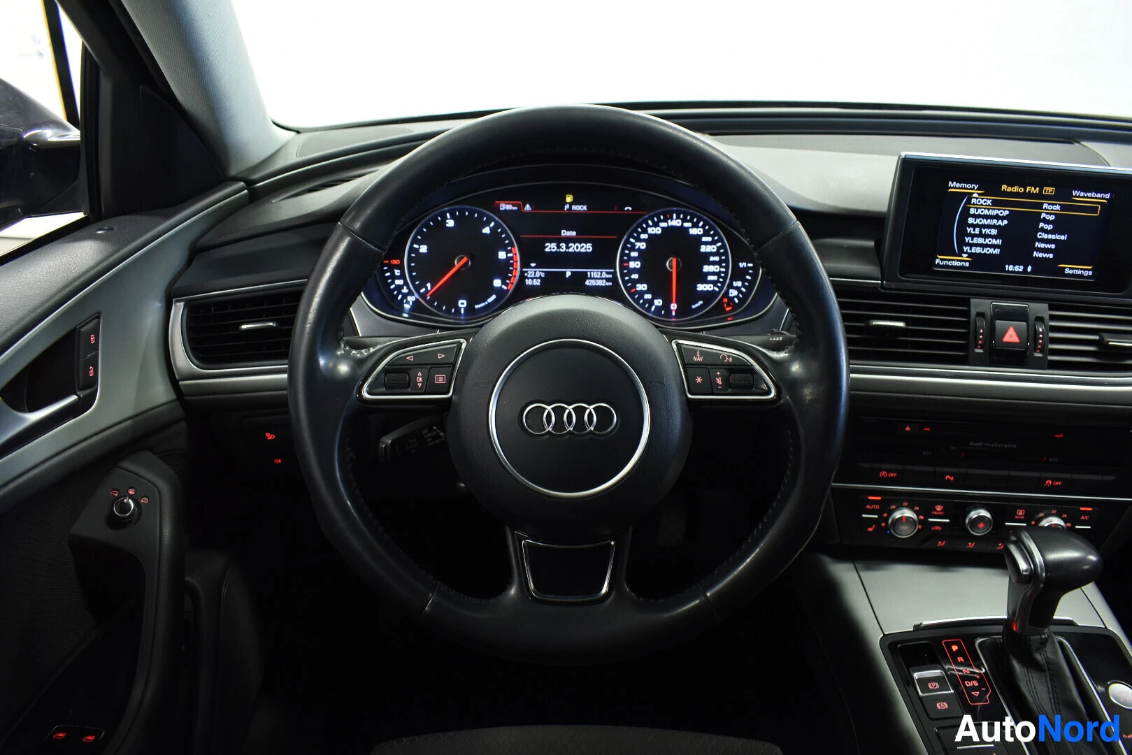 Ohjaamo: Musta monitoimiohjauspyörä, jossa on Audi-logo. Sen takana näkyy auton mittaristo ja sen oikealla puolella auton infotainment-näyttö.