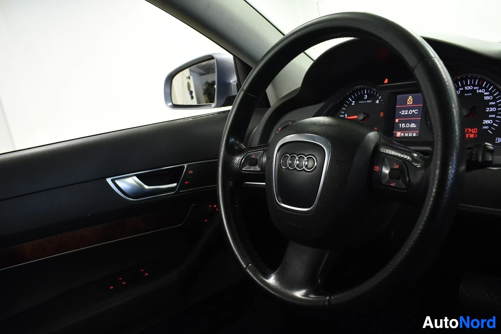 Ohjaamo: Kojelauta ja ohjauspyörä kuvattuna vasemmalta puolelta. Ohjauspyörässä on monitoimipainikkeita ja Audi-logo. Mittaristossa näkyy ulkolämpötila -22.0 °C ja kello