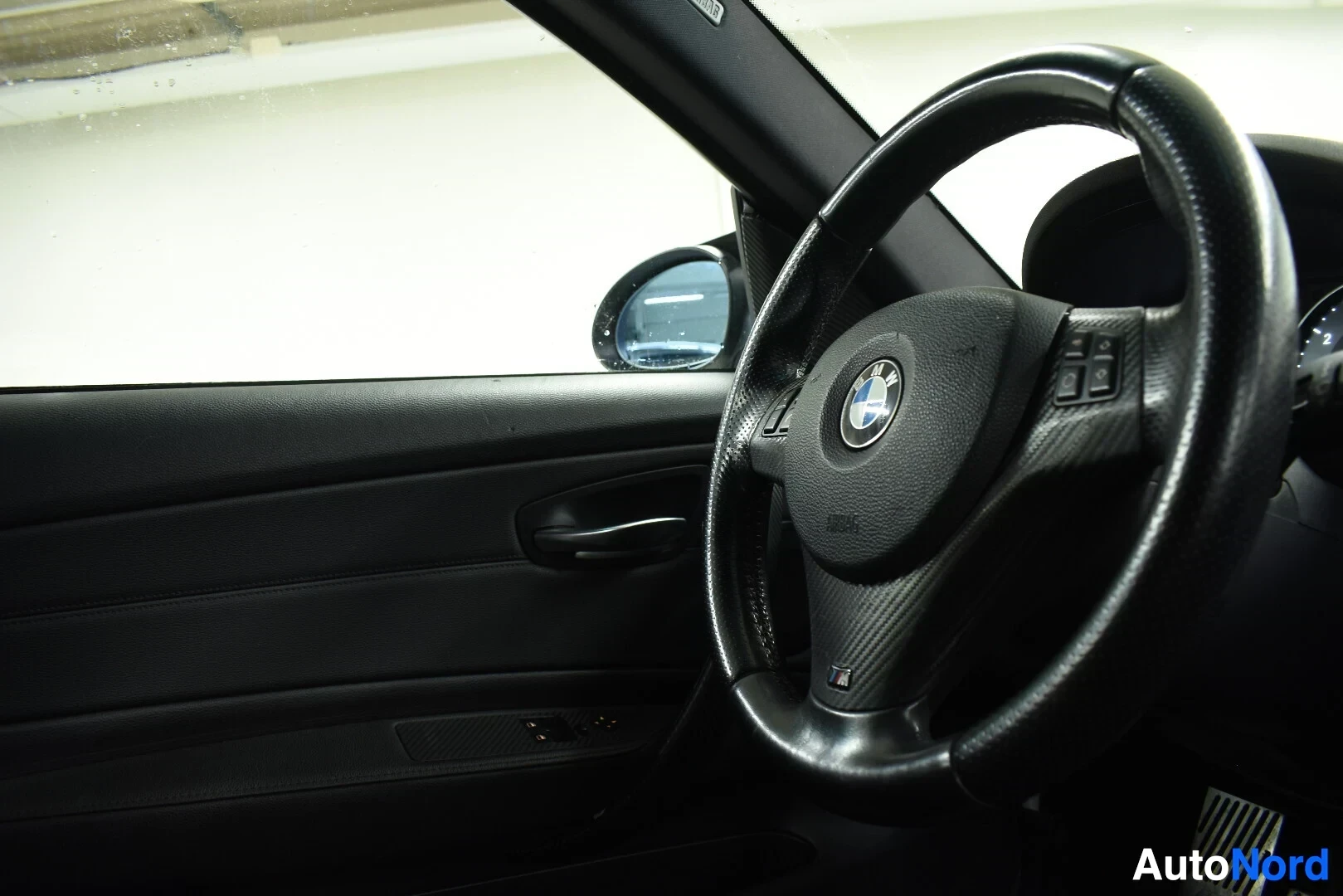 Ohjaamo: Kuvassa näkyy auton sisätila kuljettajan puolelta. Musta ohjauspyörä, jossa on BMW-logo keskellä, on kuvan etualalla. Ohjauspyörän pinnalla on hiilikuitujäljite