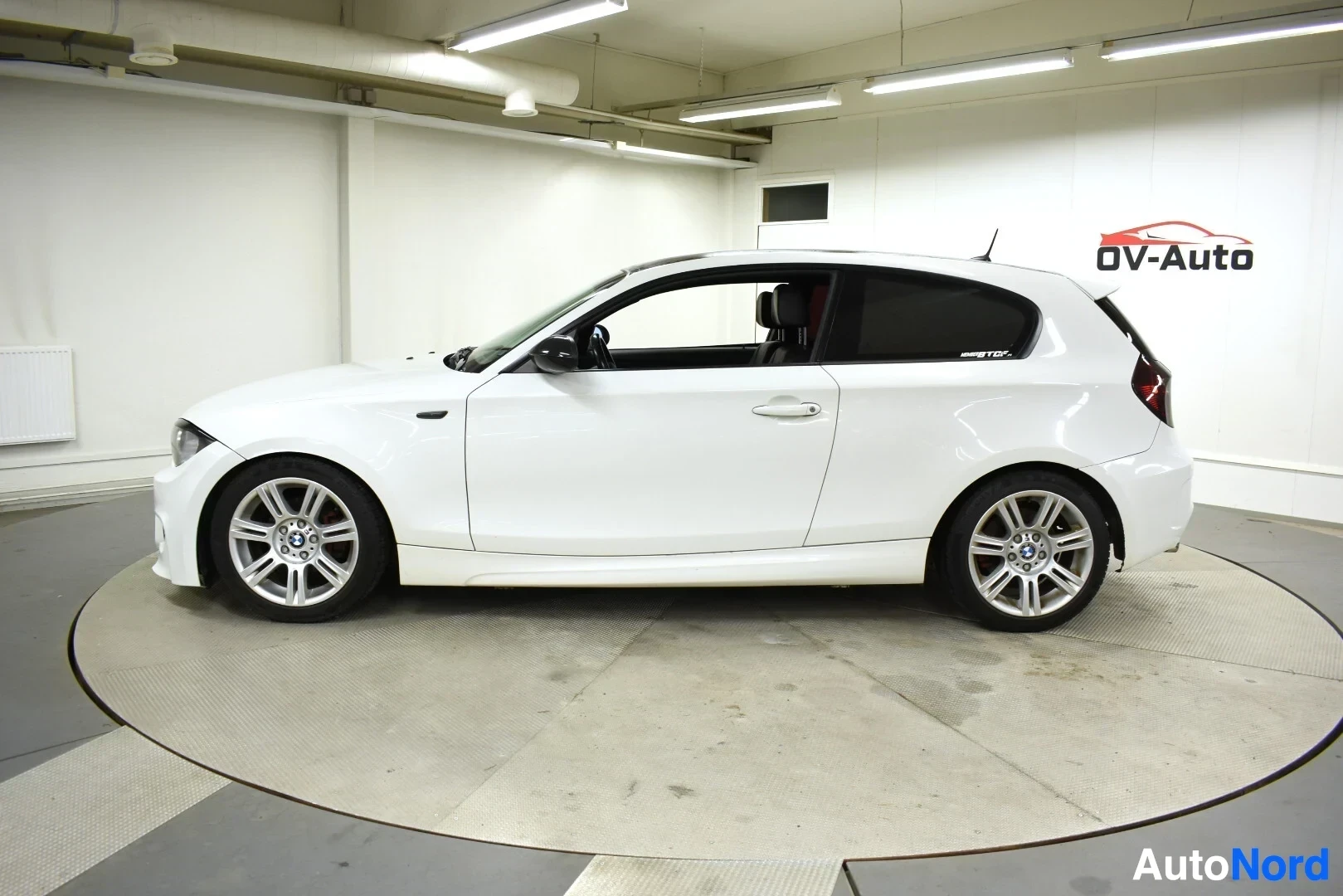 Sivulta: Valkoinen kaksiovinen BMW henkilöauto, jossa on musta katto ja vaaleat kevytmetallivanteet. Auto on kuvattu suoraan vasemmalta sivulta sisätiloissa.