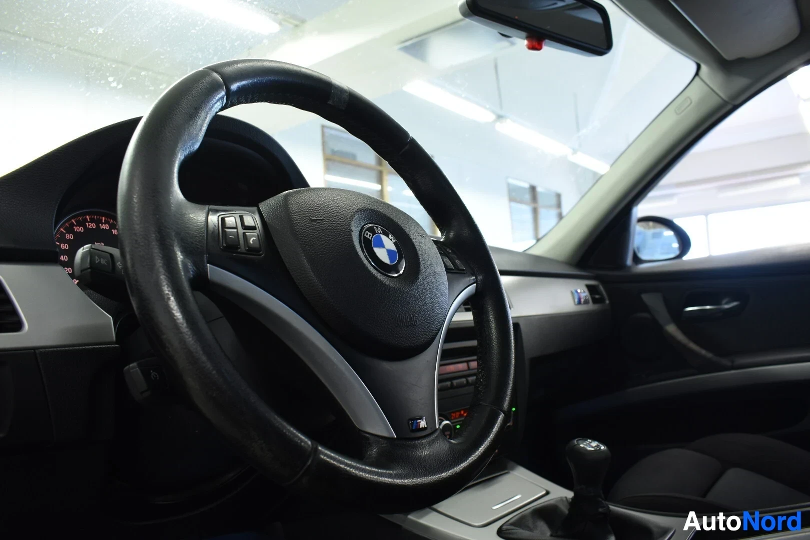 Ohjaamo: Musta nahkainen ohjauspyörä useilla painikkeilla, BMW-logo ja alareunassa M-logo. Näkyvissä on myös vaihdekeppi ja osia kojelaudasta.