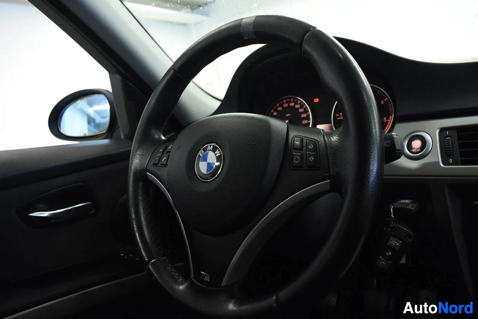 Ohjaamo: Ohjauspyörä, jossa BMW-logo, monitoimipainikkeet ja M-logo alaosassa. Mittaristo näkyy taustalla. Avaimet roikkuvat virtalukossa oikealla.