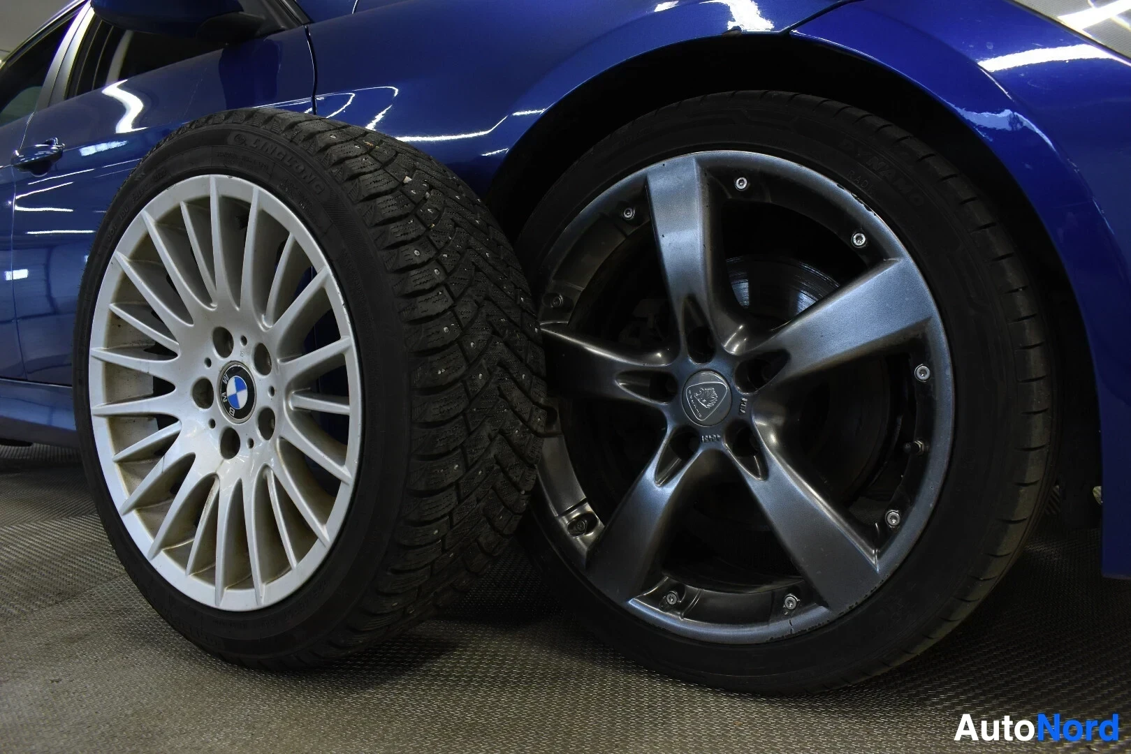 Yksityiskohta: Kaksi rengassarjaa vierekkäin auton vieressä. Vasemmalla on valkoinen kevytmetallivanne talvirenkaalla, jossa on BMW-logo keskellä. Oikealla on tummanharmaa vii