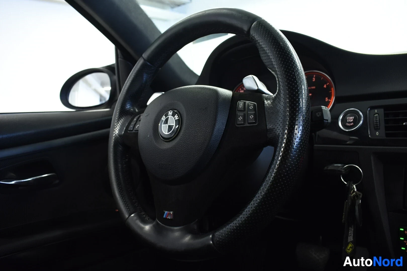 Ohjaamo: Ohjauspyörä, jossa on BMW-logo, monitoimipainikkeet ja M-logo. Näkyvissä on myös auton käynnistyspainike ja osittain mittaristo.