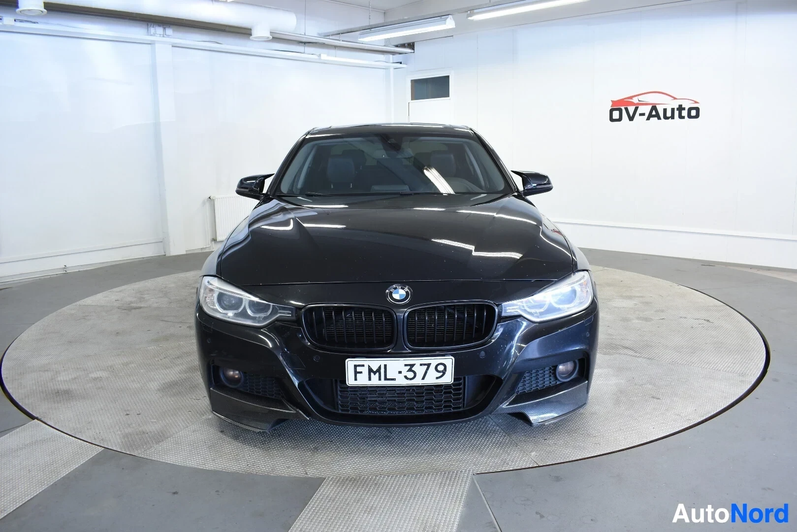Edestä: Musta sedan BMW, edestä kuvattuna pyörivällä alustalla sisätiloissa. Autossa on rekisterikilpi