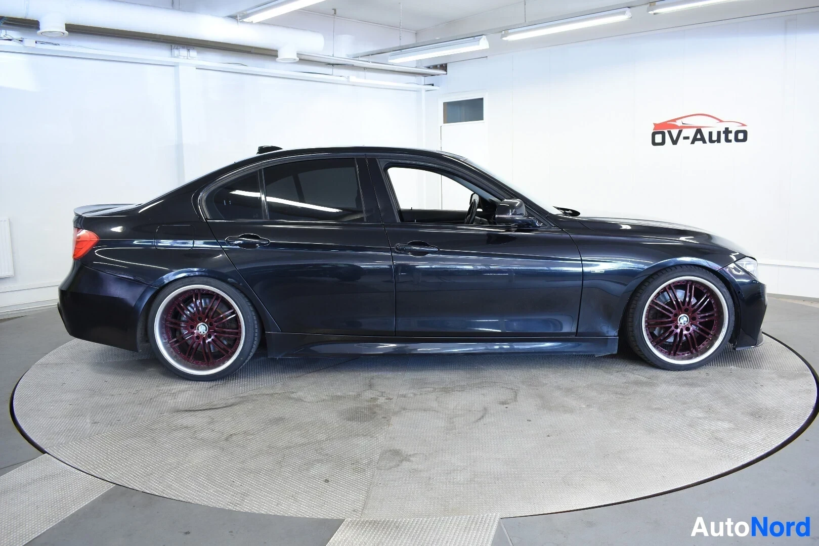 Sivulta: Musta BMW sedan vasemmalta sivulta kuvattuna. Autossa on tummanpunaiset monipuolaiset vanteet kromireunuksilla. Ikkunat ovat tummennetut.