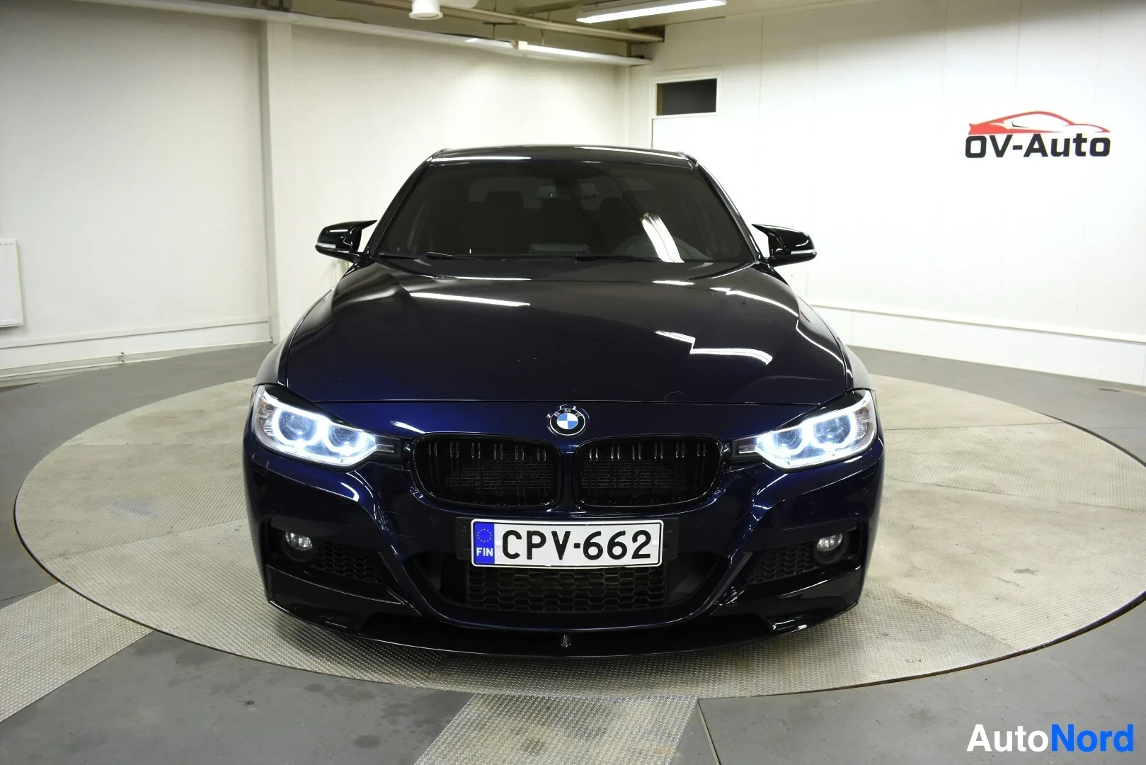 Edestä: Tummanpunainen BMW 3-sarjan sedanin etuosa edestäpäin sisätiloissa. Ajovalot ovat päällä.