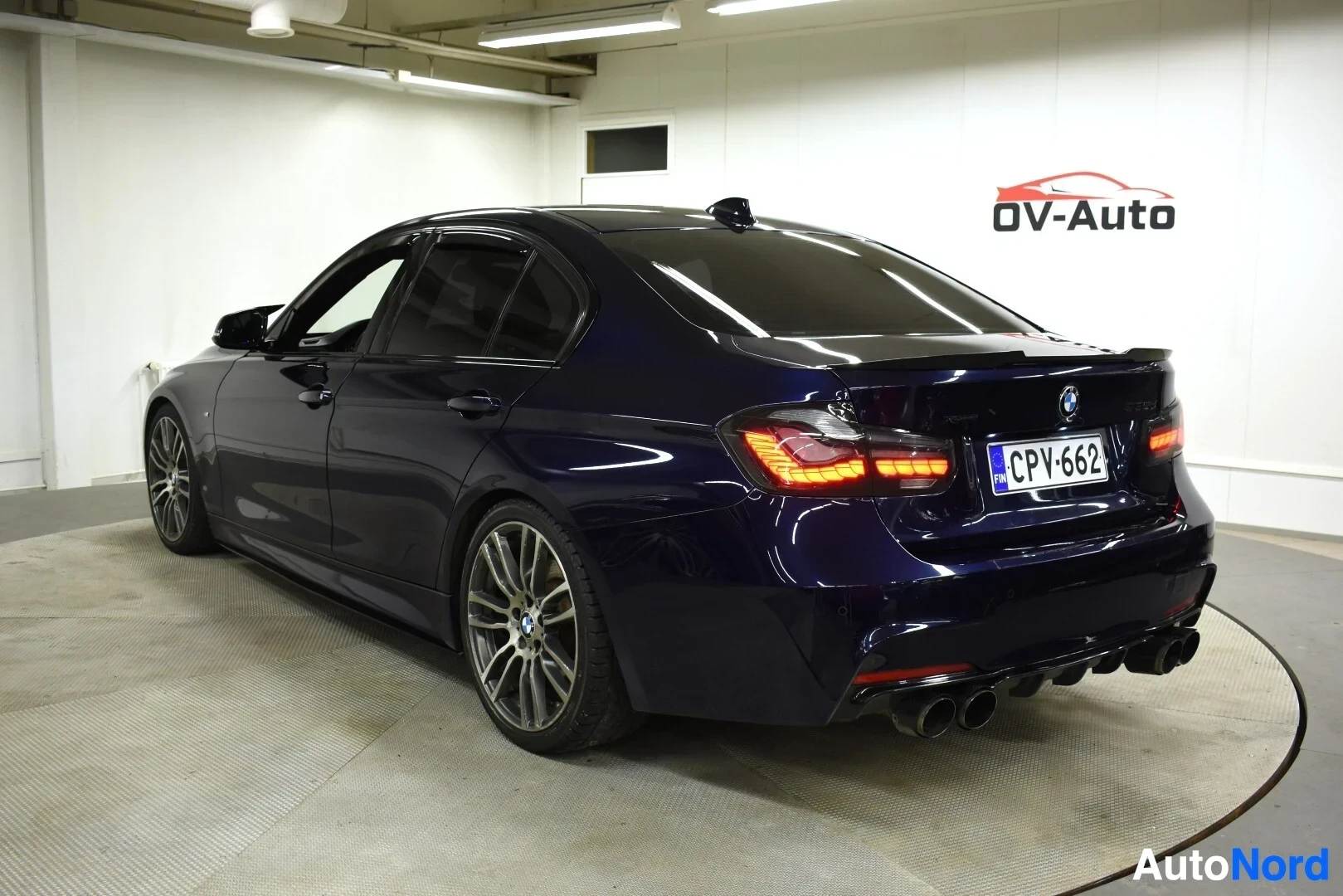 Takaviisto: Tummanpunainen BMW 3-sarjan sedan takaviistosta kuvattuna sisätiloissa. Autossa on tummennetut takalasit, neljä pakoputken ulostuloa ja monipuolaiset kevytmetal