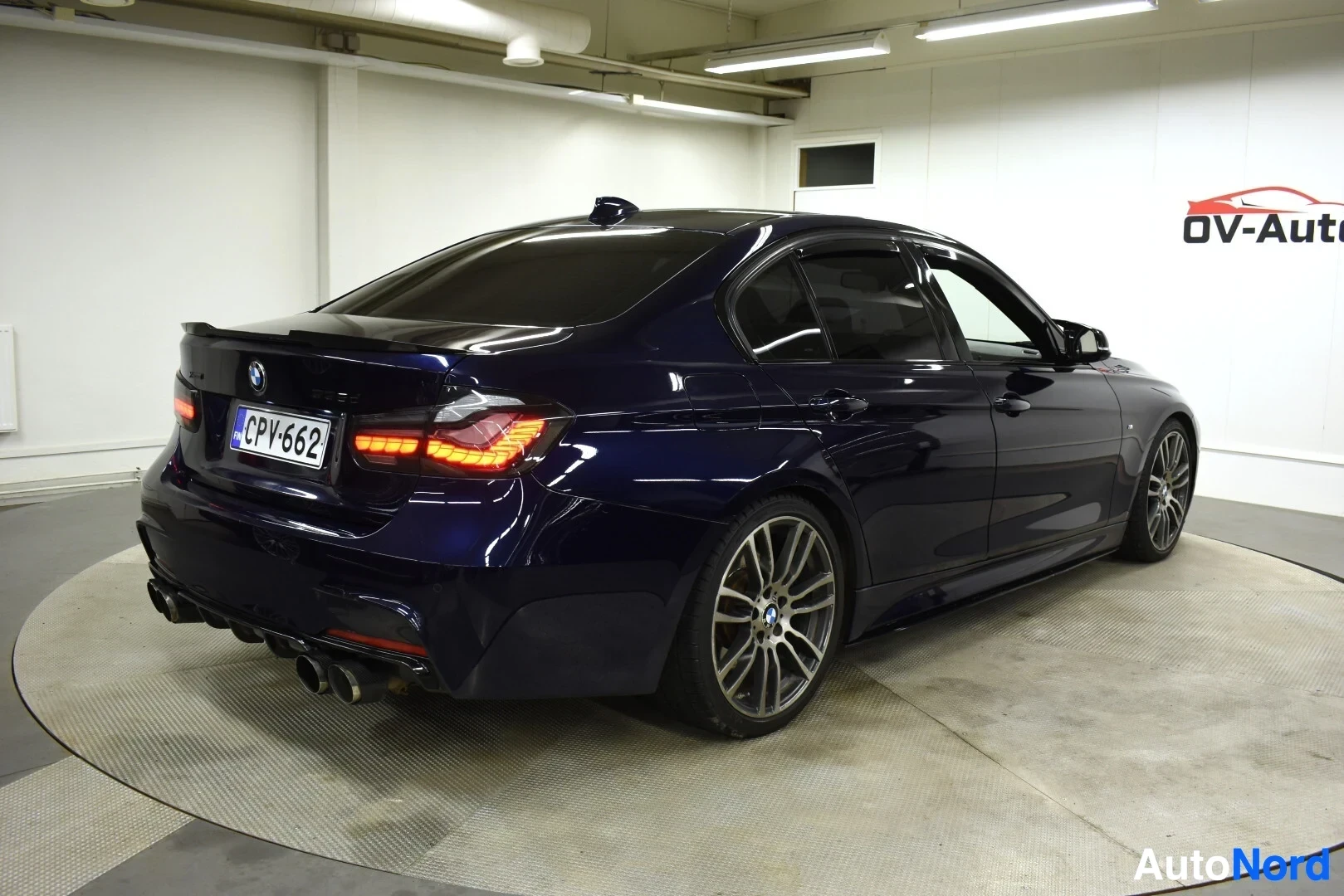 Takaviisto: Tummanpunainen BMW 3-sarjan sedan takaviistosta kuvattuna sisätiloissa. Autossa on tummennetut takalasit, neljä pakoputken ulostuloa ja monipuolaiset kevytmetal