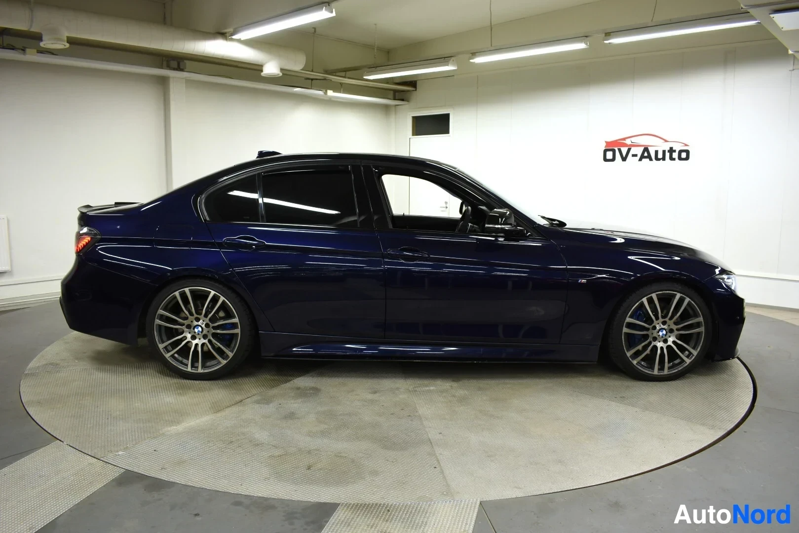 Sivulta: Tummanpunainen BMW 3-sarjan sedan sivulta kuvattuna sisätiloissa. Autossa on tummennetut takalasit ja monipuolaiset kevytmetallivanteet sinisillä jarrusatuloill