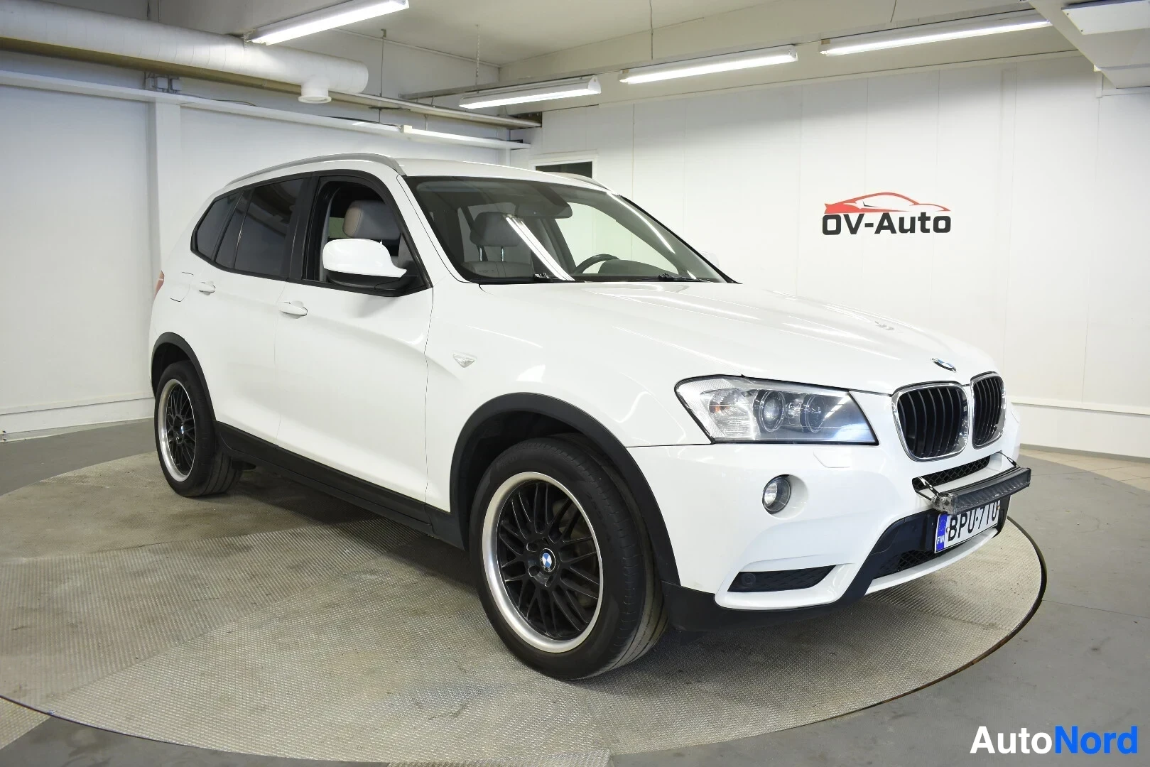 Etuviisto, nokka oikealle: Valkoinen BMW X3 -katumaasturi edestä ja vasemmalta sivulta kuvattuna sisätiloissa. Autossa on mustat monipuolavanteet, joissa on kiiltävä vannekehä.