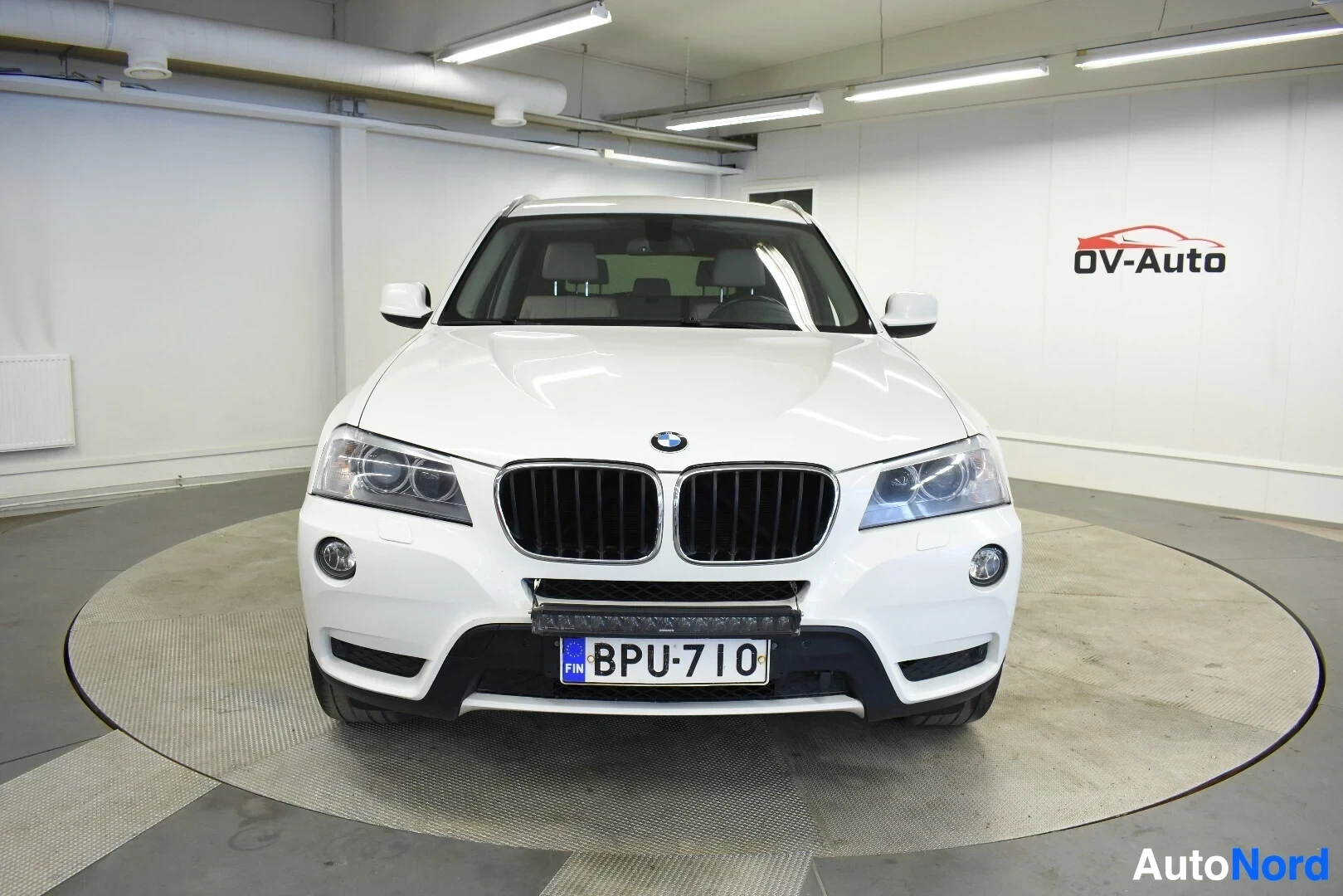 Edestä: Valkoinen BMW X3 -katumaasturi edestä päin kuvattuna sisätiloissa.