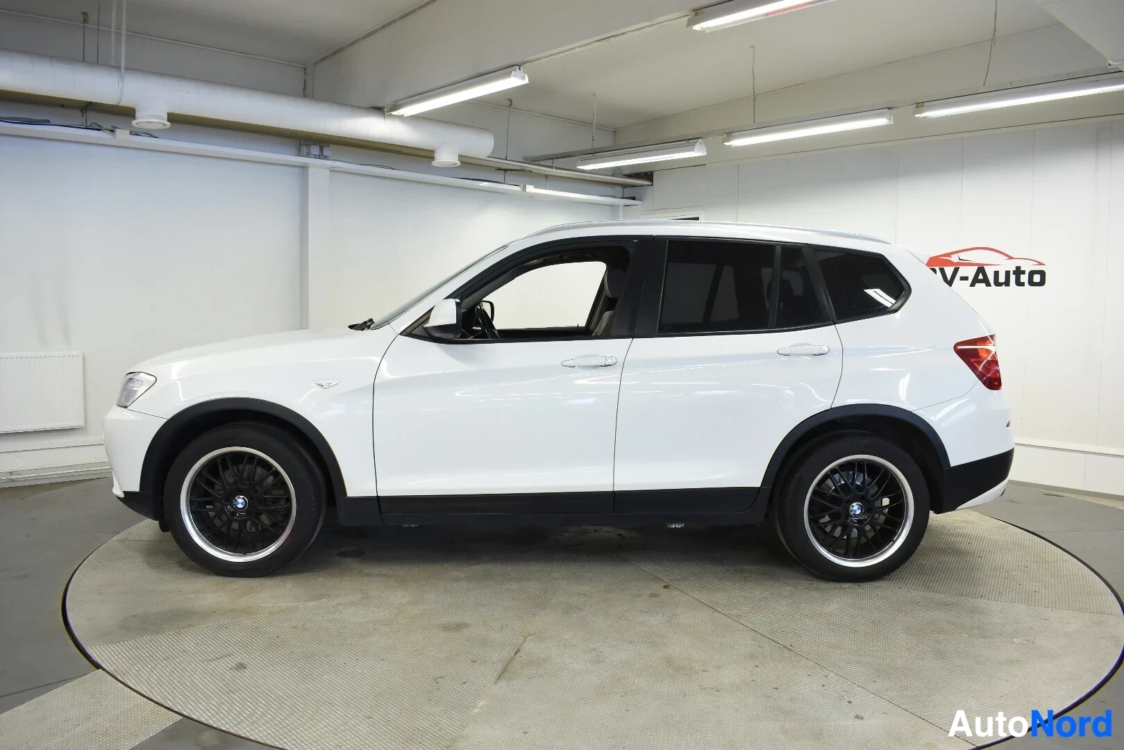 Sivulta: Valkoinen BMW X3 -katumaasturi oikealta sivulta kuvattuna sisätiloissa. Autossa on mustat monipuolavanteet, joissa on kiiltävä vannekehä.