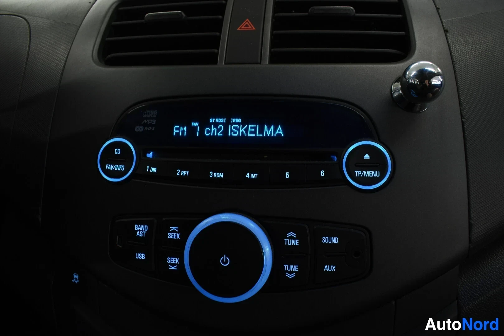 Yksityiskohta: Lähikuva auton kojelaudan radiosta, jossa on sininen taustavalo ja teksti "FM 1 ch2 ISKELMÄ".