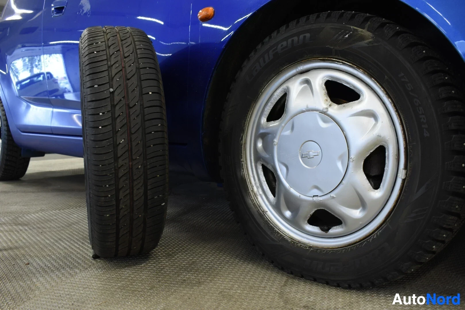 Yksityiskohta: Kuva auton kyljestä, jossa näkyy auton alle asennettu nastarengas ja auton vieressä pystyssä seisova nastarengas. Auton alle asennetussa renkaassa on Chevrolet-