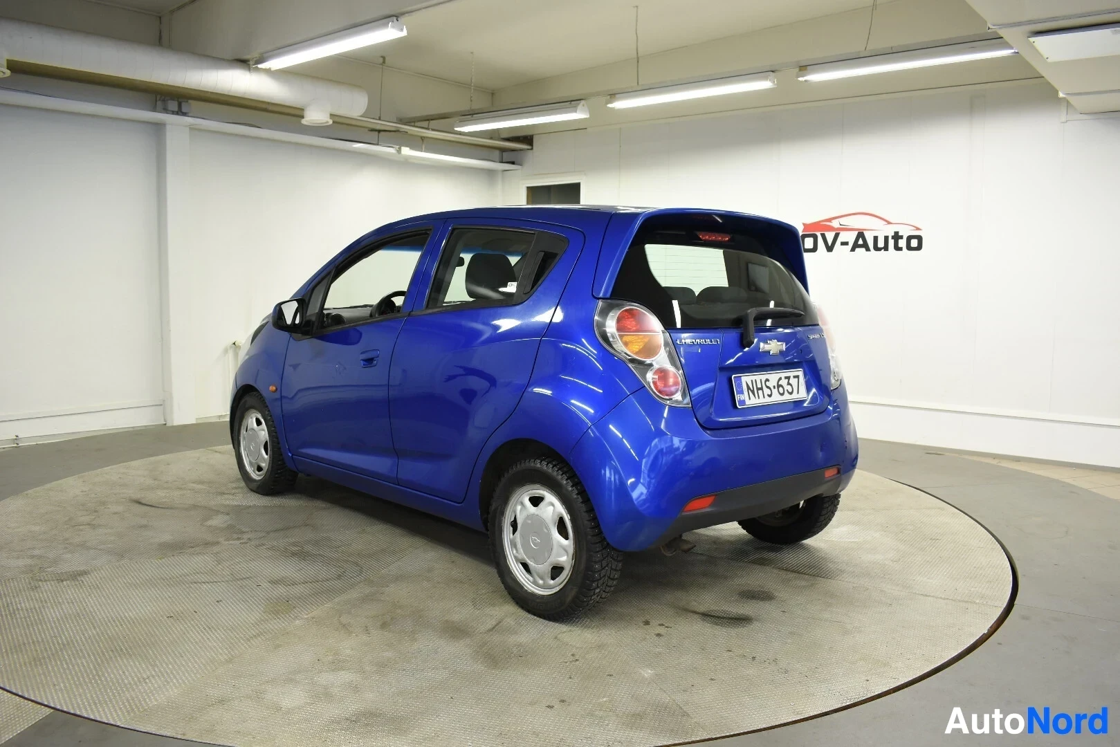 Takaviisto: Sininen hatchback-auto pyörivällä alustalla, näkyvissä takaa oikealta puolelta. Autossa on valkoiset vanteet ja takarekisterikilpi ""