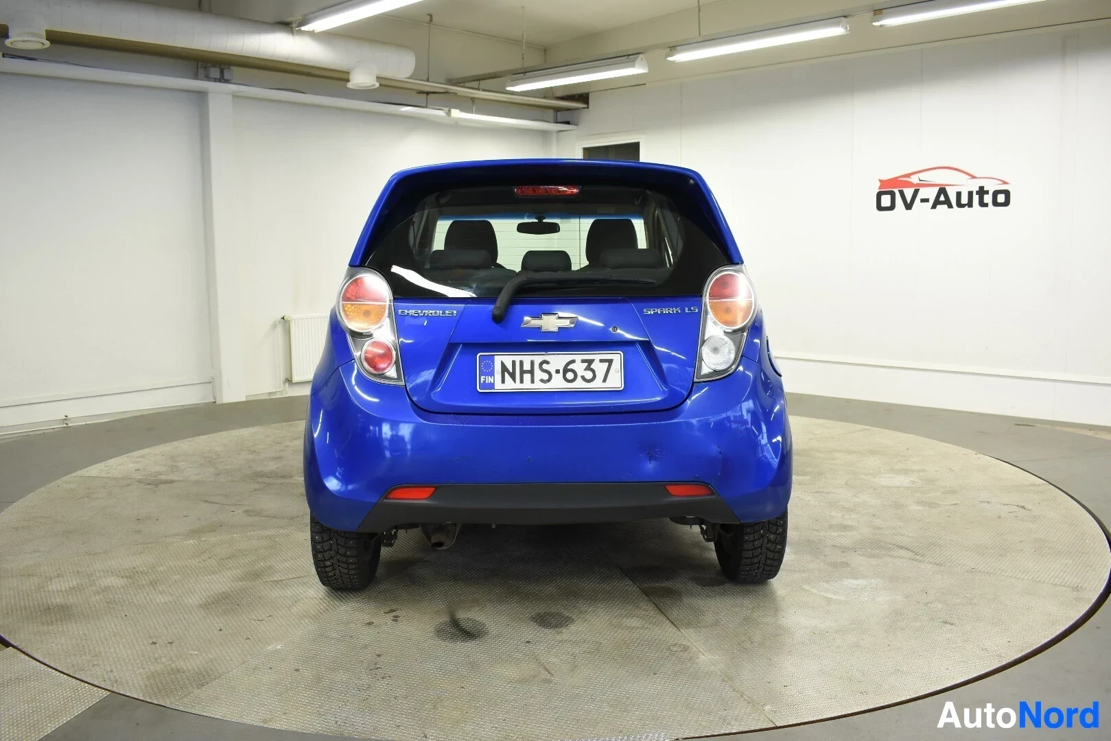 Takaa: Sininen hatchback-auto takaa päin pyörivällä alustalla. Takarekisterikilpi "" ja 'Chevrolet' sekä 'Spark LS' -merkit ovat näkyvissä
