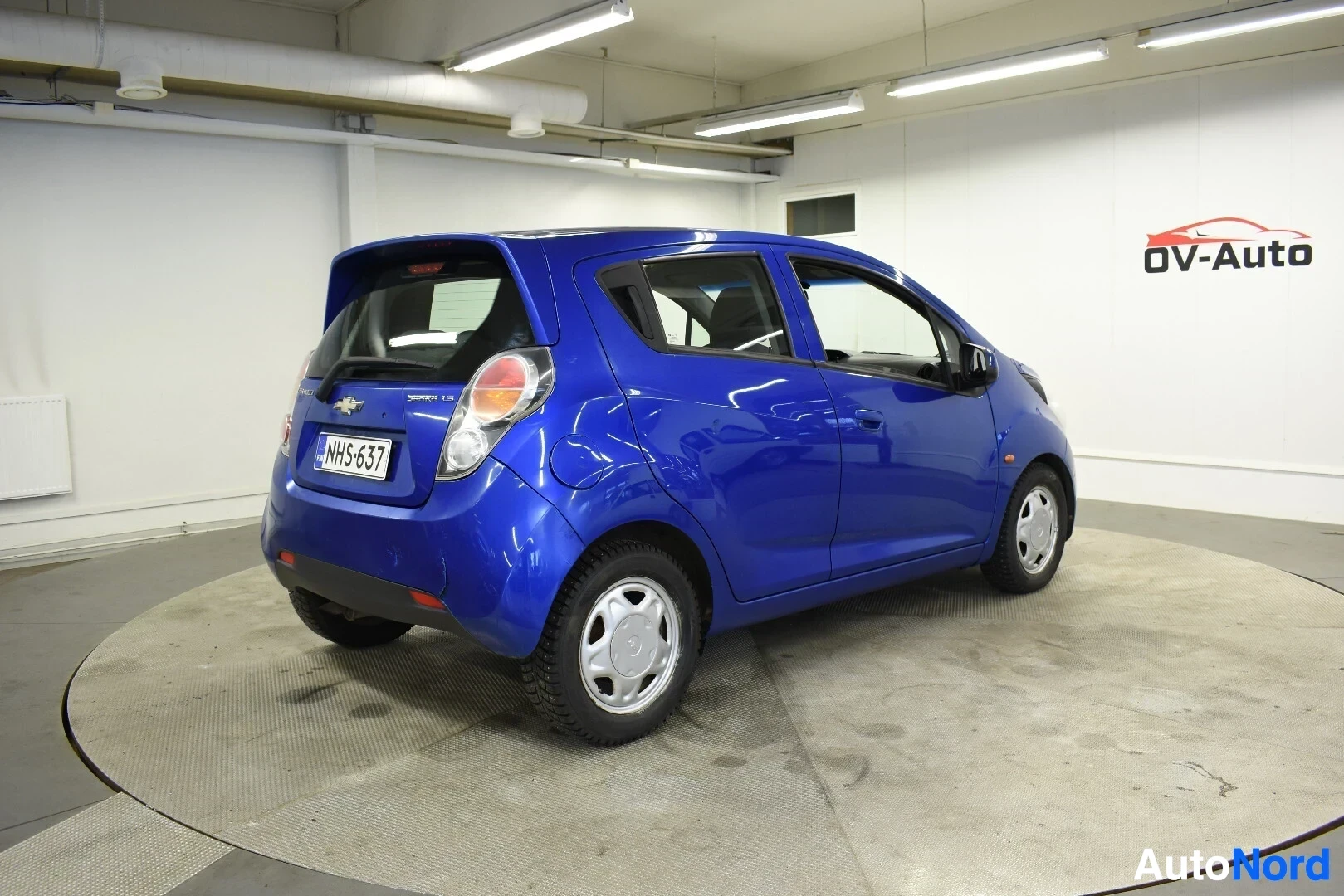 Takaviisto: Sininen hatchback-auto pyörivällä alustalla, näkyvissä takaa vasemmalta puolelta. Autossa on valkoiset vanteet ja takarekisterikilpi ""