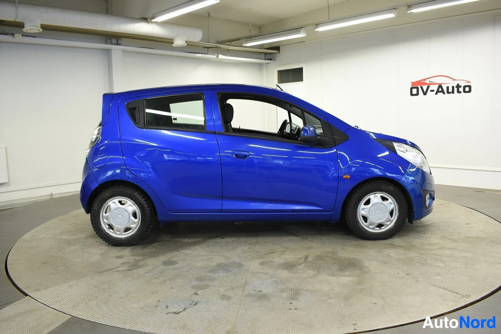 Sivulta: Sininen hatchback-auto vasemmalta sivulta pyörivällä alustalla. Autossa on valkoiset vanteet.