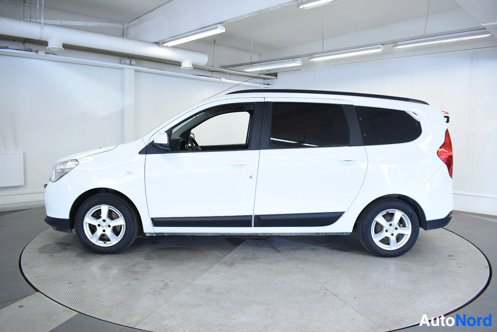 Sivulta: Valkoinen tila-auto kuvattuna suoraan oikealta sivulta. Autossa on kattokaiteet ja alumiinivanteet.