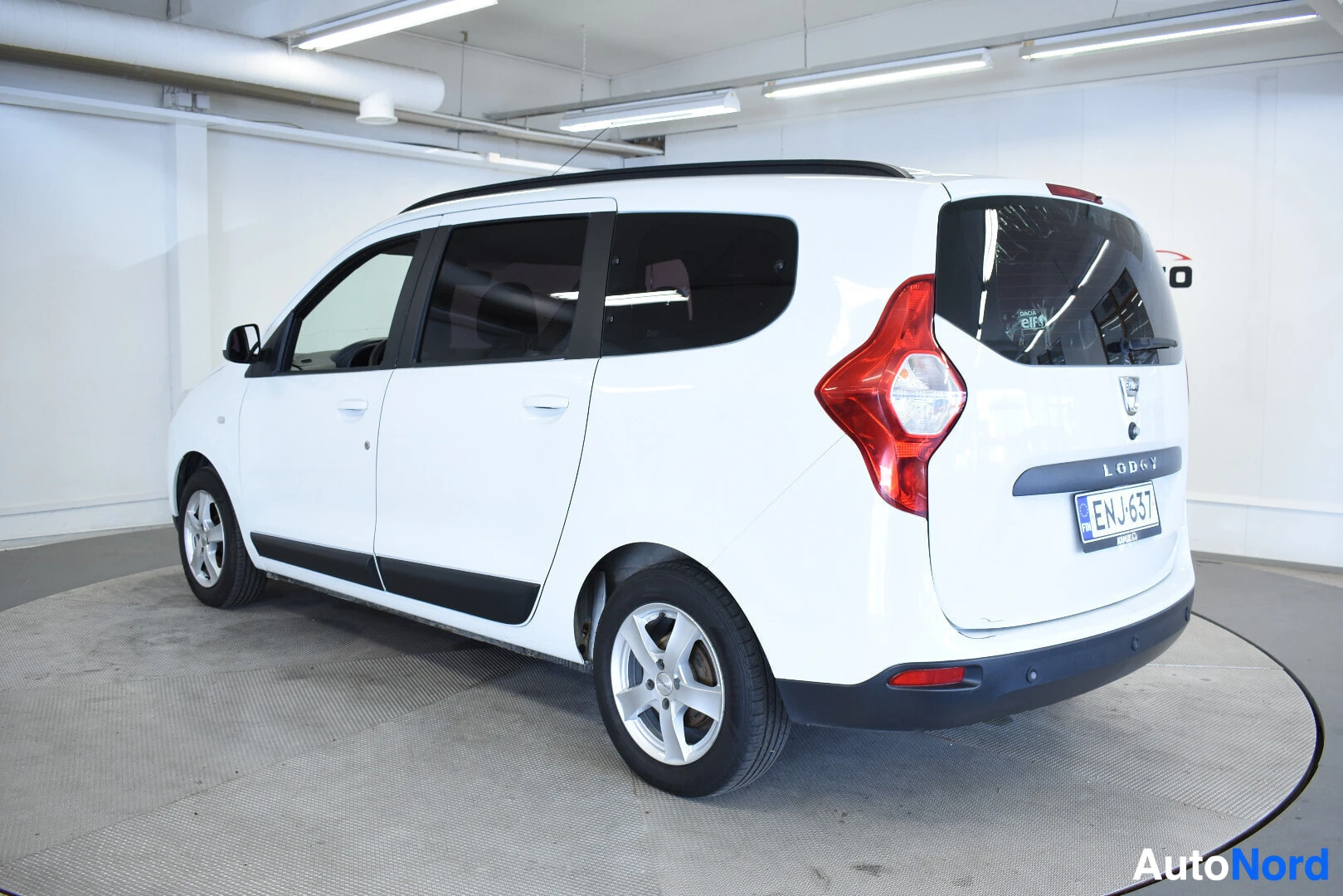 Takaviisto: Valkoinen Dacia Lodgy henkilöauto kuvattuna takaviistosta oikealta sisätiloissa. Autossa on kattokiskot ja suomalainen rekisterikilpi ""
