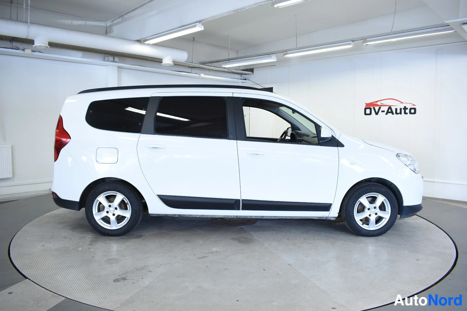 Sivulta: Valkoinen Dacia Lodgy henkilöauto kuvattuna suoraan sivulta sisätiloissa. Autossa on kattokiskot.