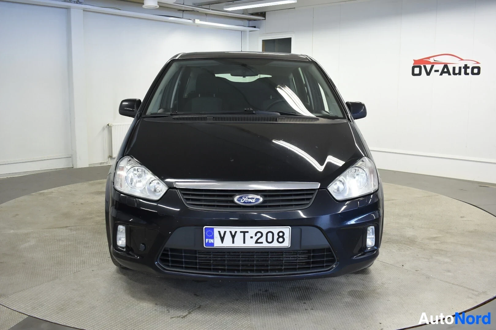 Edestä: Musta henkilöauto pysäköitynä valaistuun sisätilaan, kuvattuna edestä. Autossa on Ford-logo ja rekisterikilpi