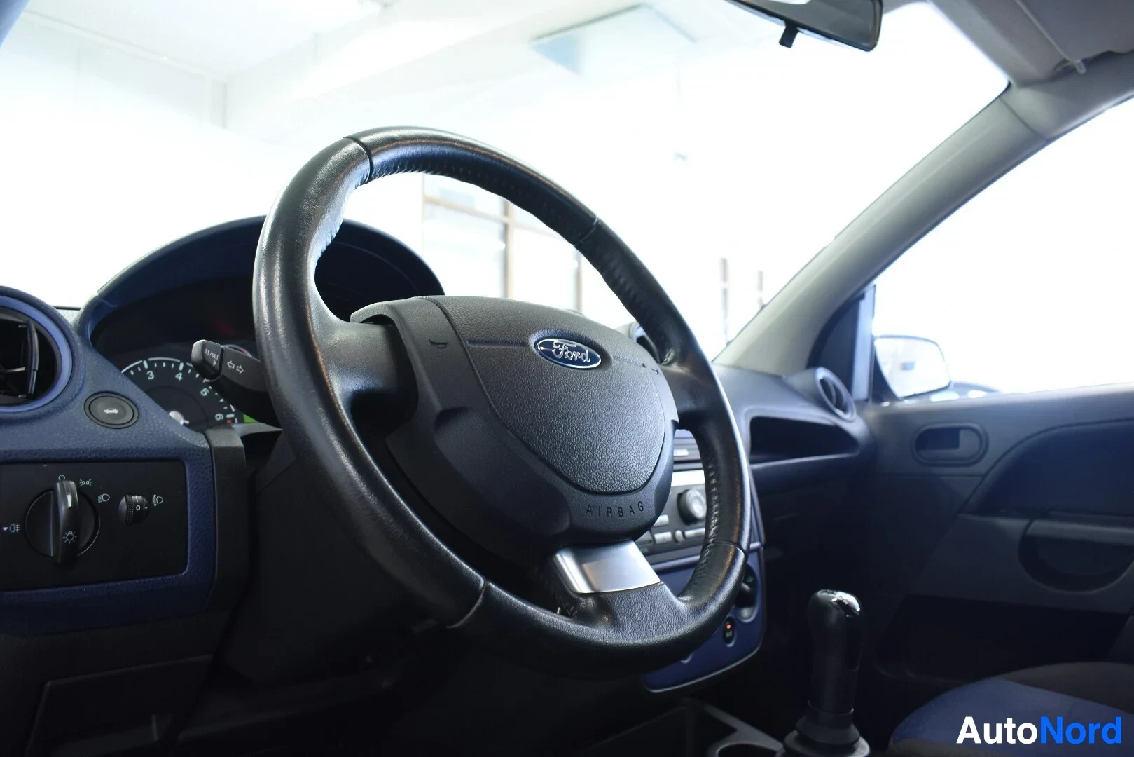 Ohjaamo: Nahkapäällysteinen ohjauspyörä, jossa Ford-logo ja sana AIRBAG keskellä, näkyy läheltä. Kojelaudan vasen puoli ja radion painikkeet näkyvät taustalla. Manuaaliv