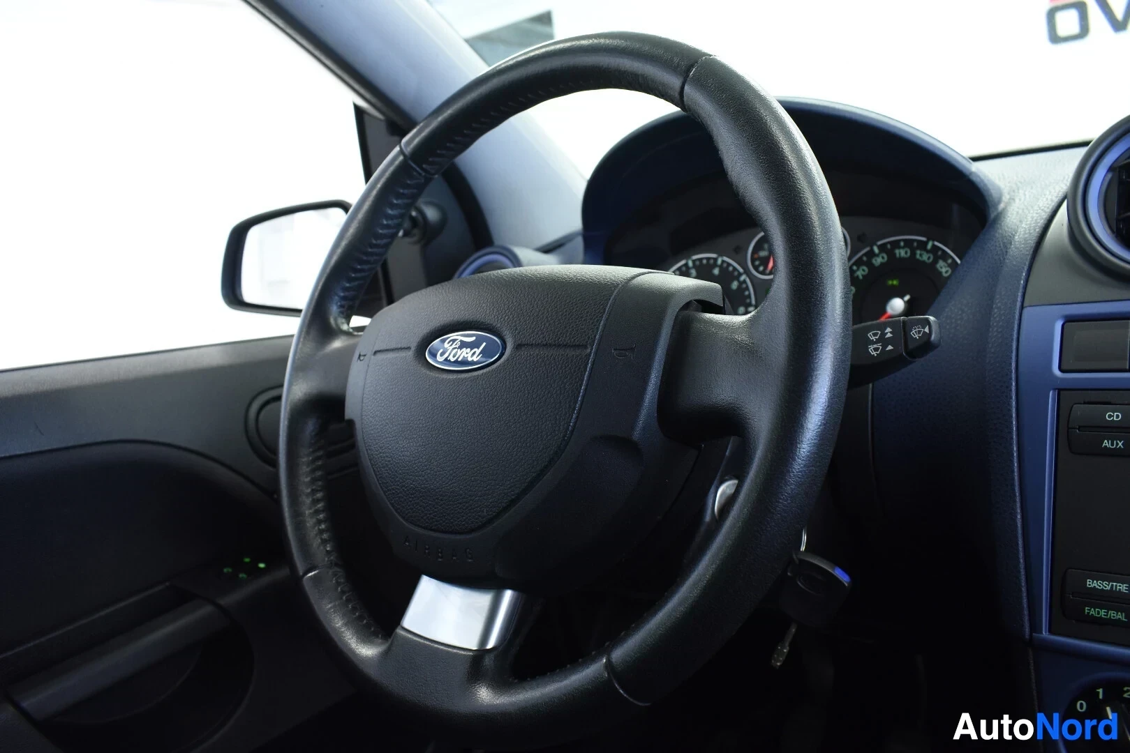 Yksityiskohta: Lähikuva ohjauspyörästä, jossa on Ford-logo ja harmaa alareuna. Ohjauspyörän oikealla puolella on virtalukko, johon on asetettu auton avaimet. Avainnipussa on k