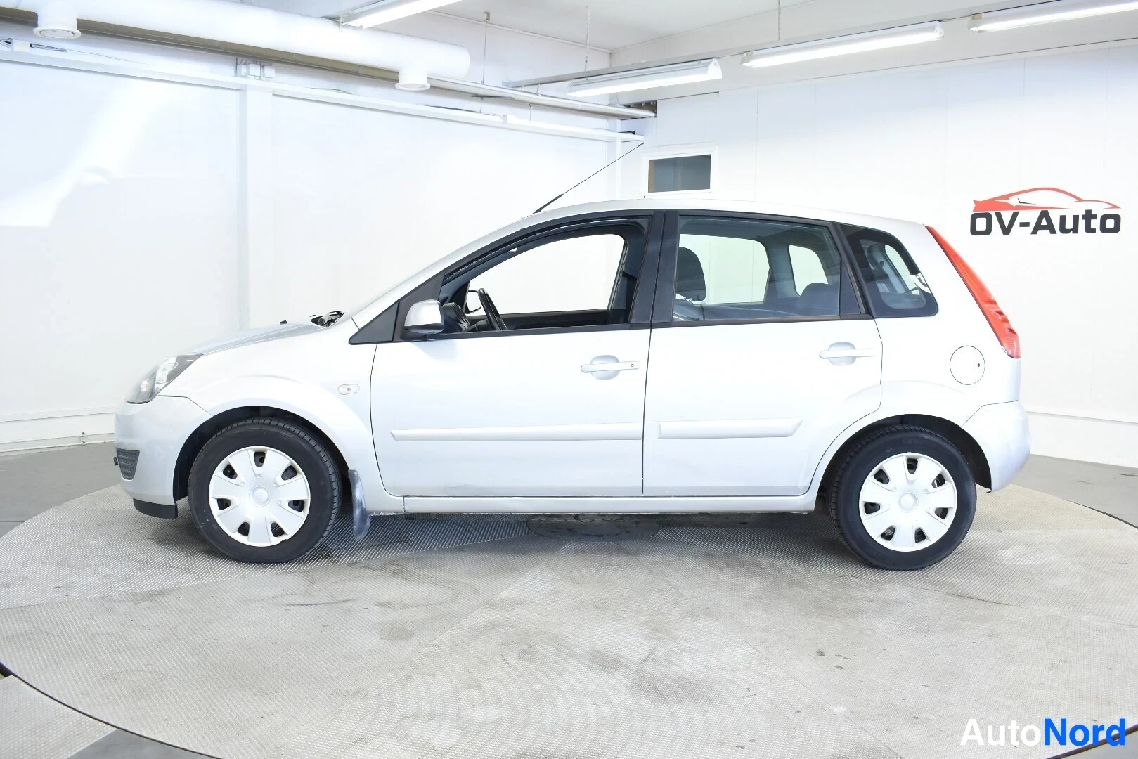 Sivulta: Hopeanvärinen henkilöauto vasemmalta sivulta kuvattuna. Autossa on valkoiset vanteiden pölykapselit.