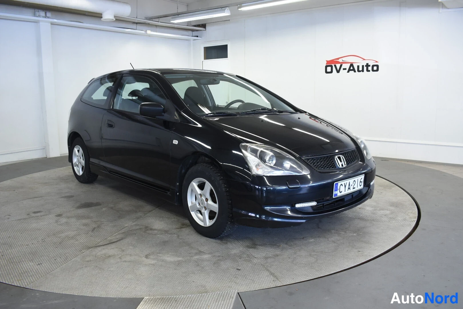 Etuviisto, nokka oikealle: Mustan Honda Civigin etukulmakuva autotallissa, rekisterikilpi, hatchback-korinen kaksiovinen auto pyörivällä alustalla.