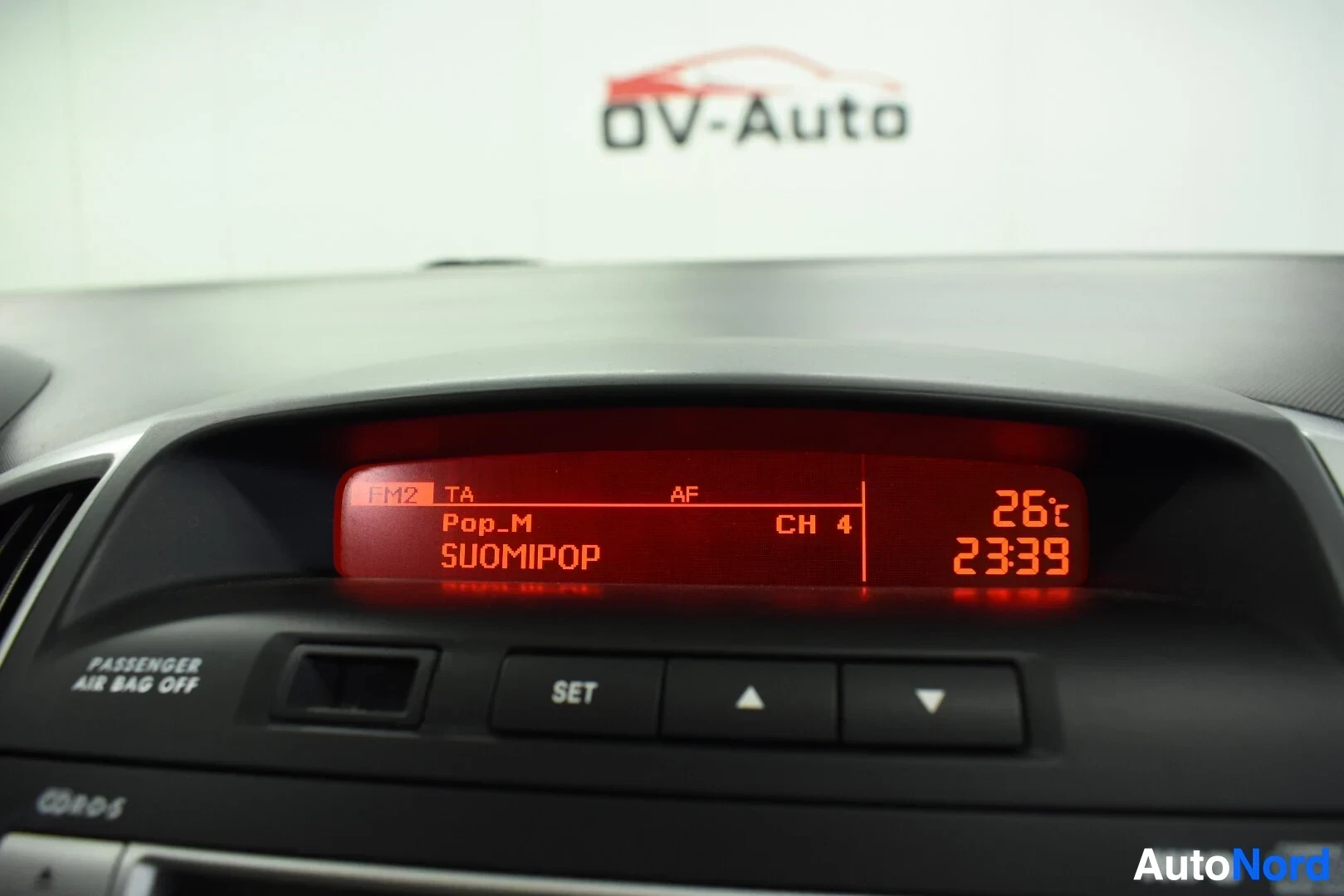 Yksityiskohta: Kojelaudan päällä oleva näyttö, jossa näkyy tekstiä "FM2 TA Pop.M SUOMIPOP" ja oikealla "26c 23:39". Näytön alla on "PASSENGER AIR BAG OFF" -merkkivalo ja paini