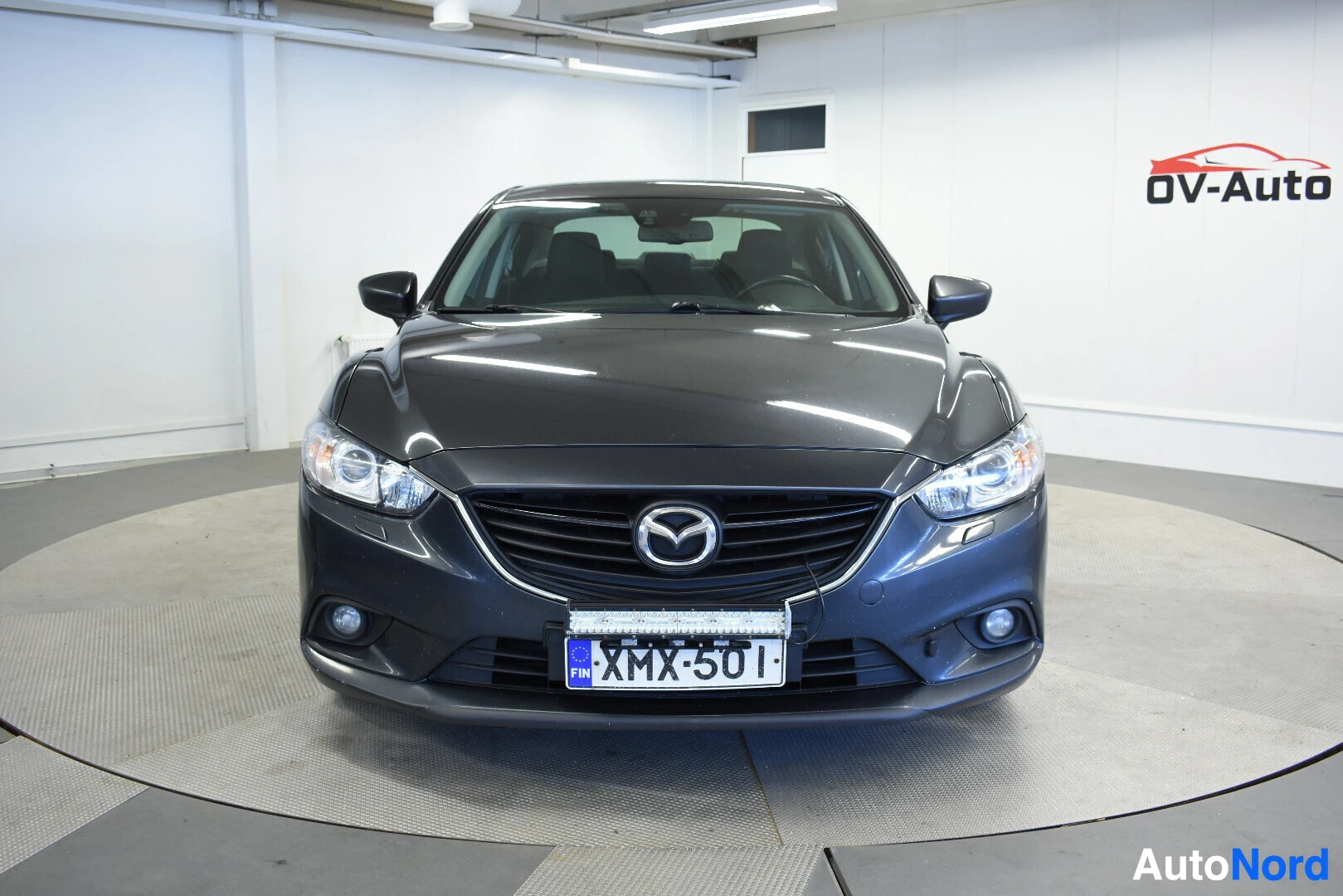 Edestä: Tummanharmaa Mazda 6 sedan suoraan edestä kuvattuna. Rekisterikilpi on ja sen yläpuolella on lisävalo. Auton edessä näkyvät ajovalot ja maski. Taustalla