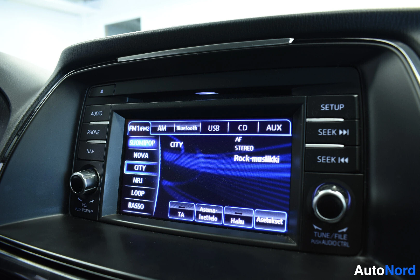 Yksityiskohta: Lähikuva auton infotainment-näytöstä, jossa on näkyvissä radion kanavalista ja yläpalkissa valittuina välilehdet FM1/RM2, AM, Bluetooth, USB, CD ja AUX. Näytöll