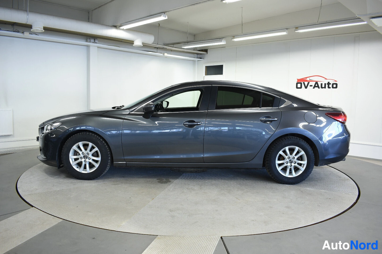 Sivulta: Tummanharmaa Mazda 6 sedan oikealta sivulta kuvattuna. Autossa on talvirenkaat. Taustalla on valkoinen halli, jossa OV-Auto logo.