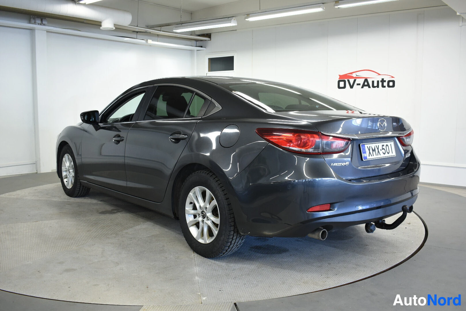 Takaviisto: Harmaa Mazda 6 sedan talvirenkailla, jossa vetokoukku, kuvattuna takavasemmalta nurin autoliikkeen tiloissa.