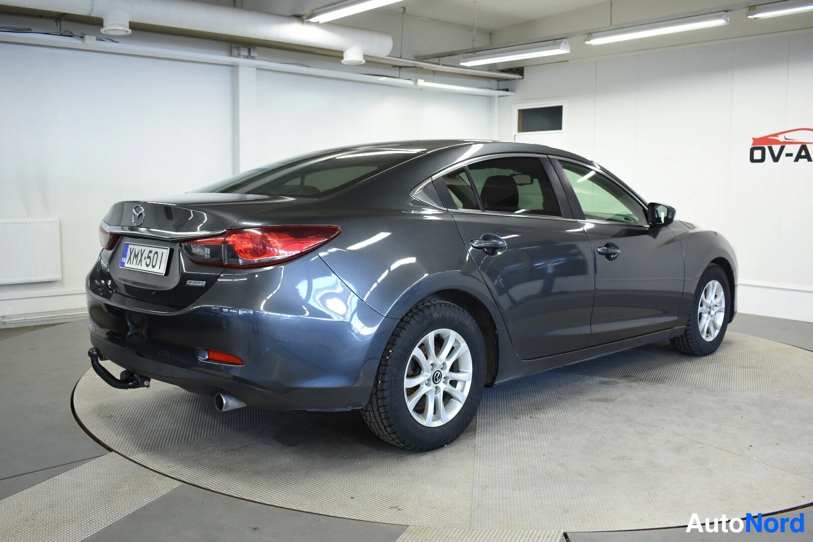 Takaviisto: Harmaa Mazda 6 sedan talvirenkailla, jossa vetokoukku, kuvattuna takaoikealta autoliikkeen tiloissa.