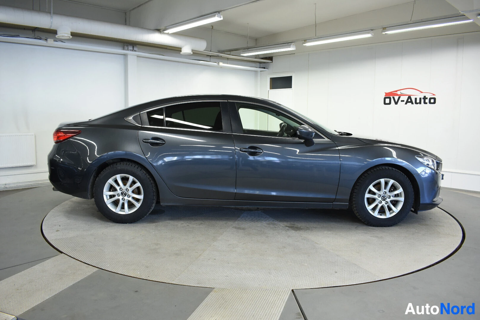 Sivulta: Harmaa Mazda 6 sedan talvirenkailla kuvattuna oikealta sivulta autoliikkeen tiloissa.