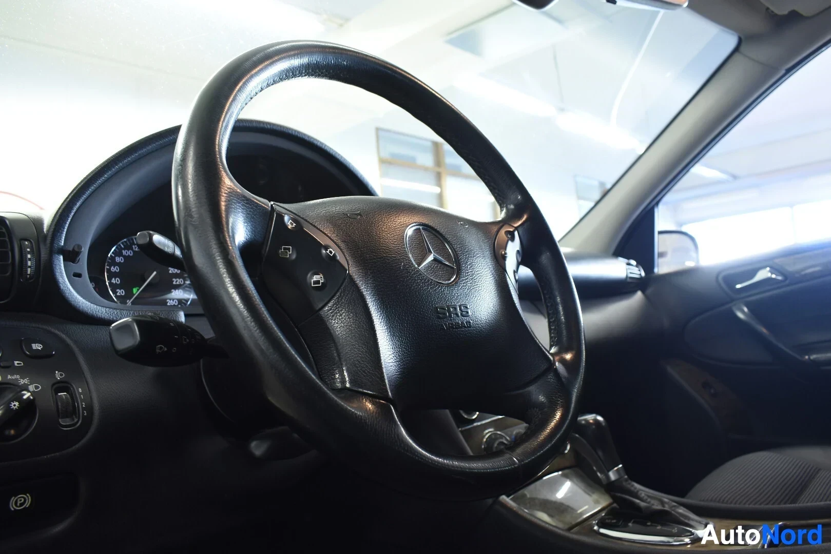 Ohjaamo: Kuljettajan puoleinen ohjauspyörä ja osa kojelautaa näkyvät vinosti kuljettajan puolelta kuvattuna. Ohjauspyörässä on Mercedes-Benzin logo ja painikkeita.