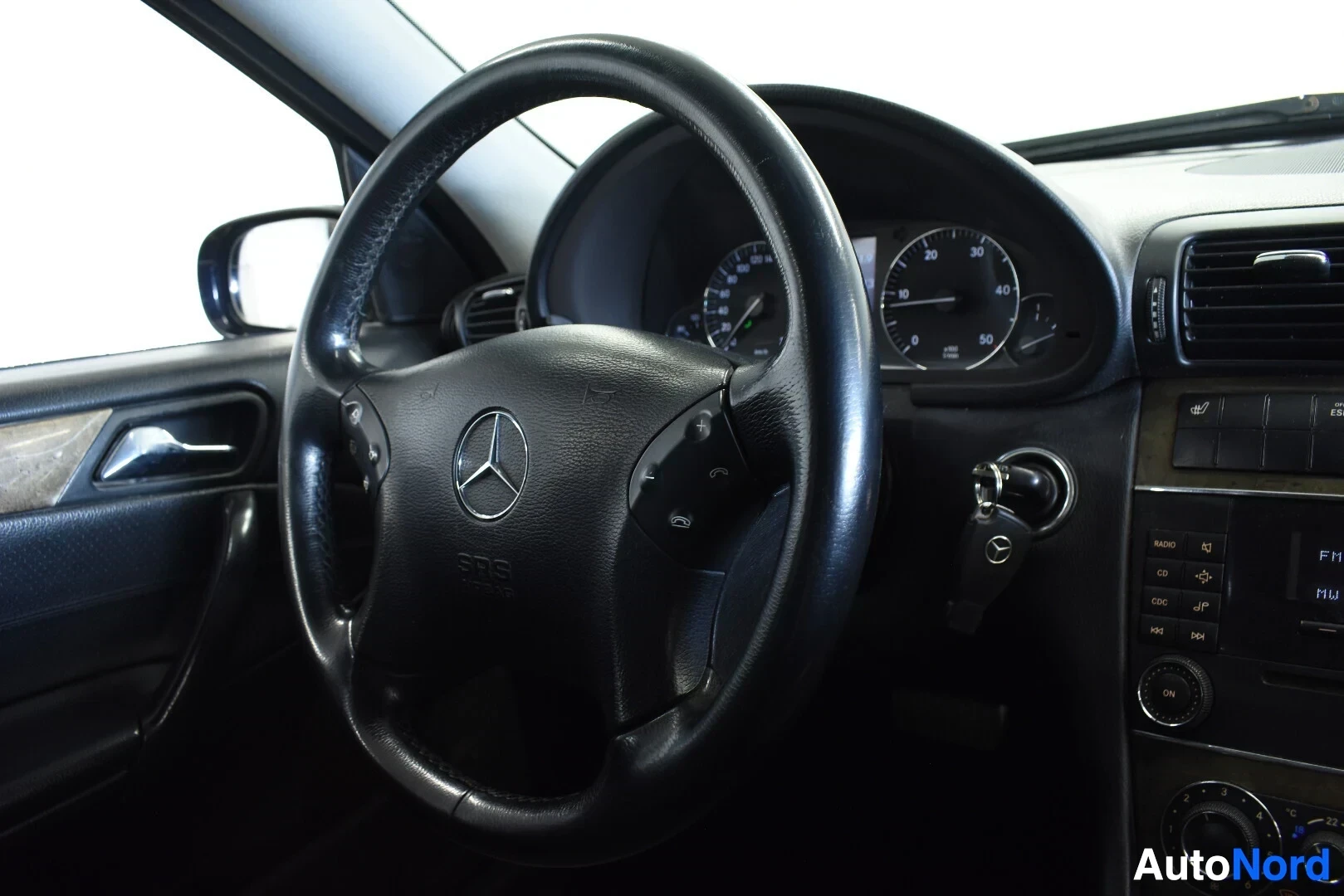 Ohjaamo: Ohjauspyörä ja osa kojelautaa kuvattuna hieman yläkulmasta kuljettajan puolelta. Ohjauspyörässä on Mercedes-Benzin logo ja multimediapainikkeita. Avaimet ovat v