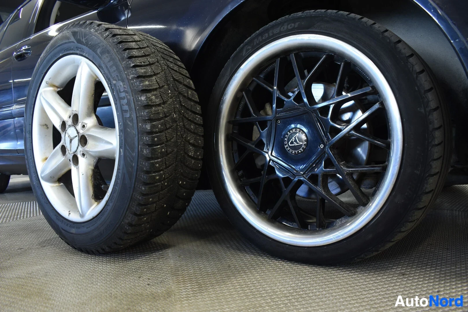 Yksityiskohta: Kuva auton pyöräkotelon edessä seisovasta lumirenkaasta ja autoon kiinnitetystä renkaasta. Molemmissa on kevytmetallivanteet.