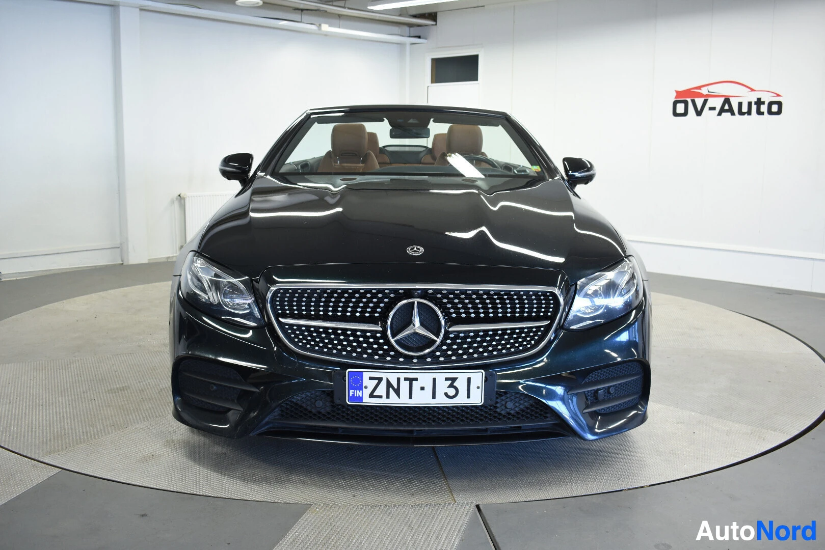 Edestä: Tummanvihreä Mercedes-Benz E-sarjan avoauto edestäpäin kuvattuna, katto alhaalla. Autossa on vaaleanruskea sisustus ja rekisterikilpi on selkeästi näkyv