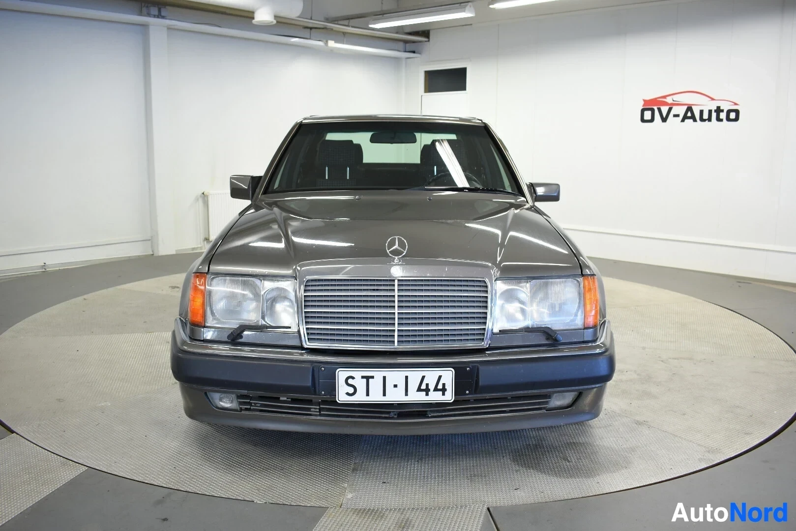 Edestä: Mercedes-Benz E 500e auton etupää on kuvattu edestä, seisottaa vaaleanharmaalla pyörivällä alustalla. Auton rekisterikilvessä lukee ""