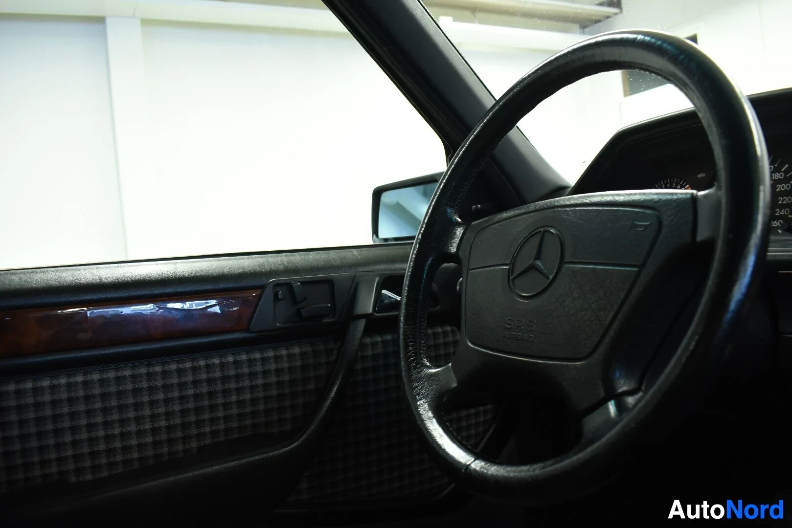 Ohjaamo: Ohjauspyörä, jossa on Mercedes-Benz-logo, ja avoin auton ovi, jossa on ruudullinen kangasverhoilu ja puujäljitelmäkoriste.