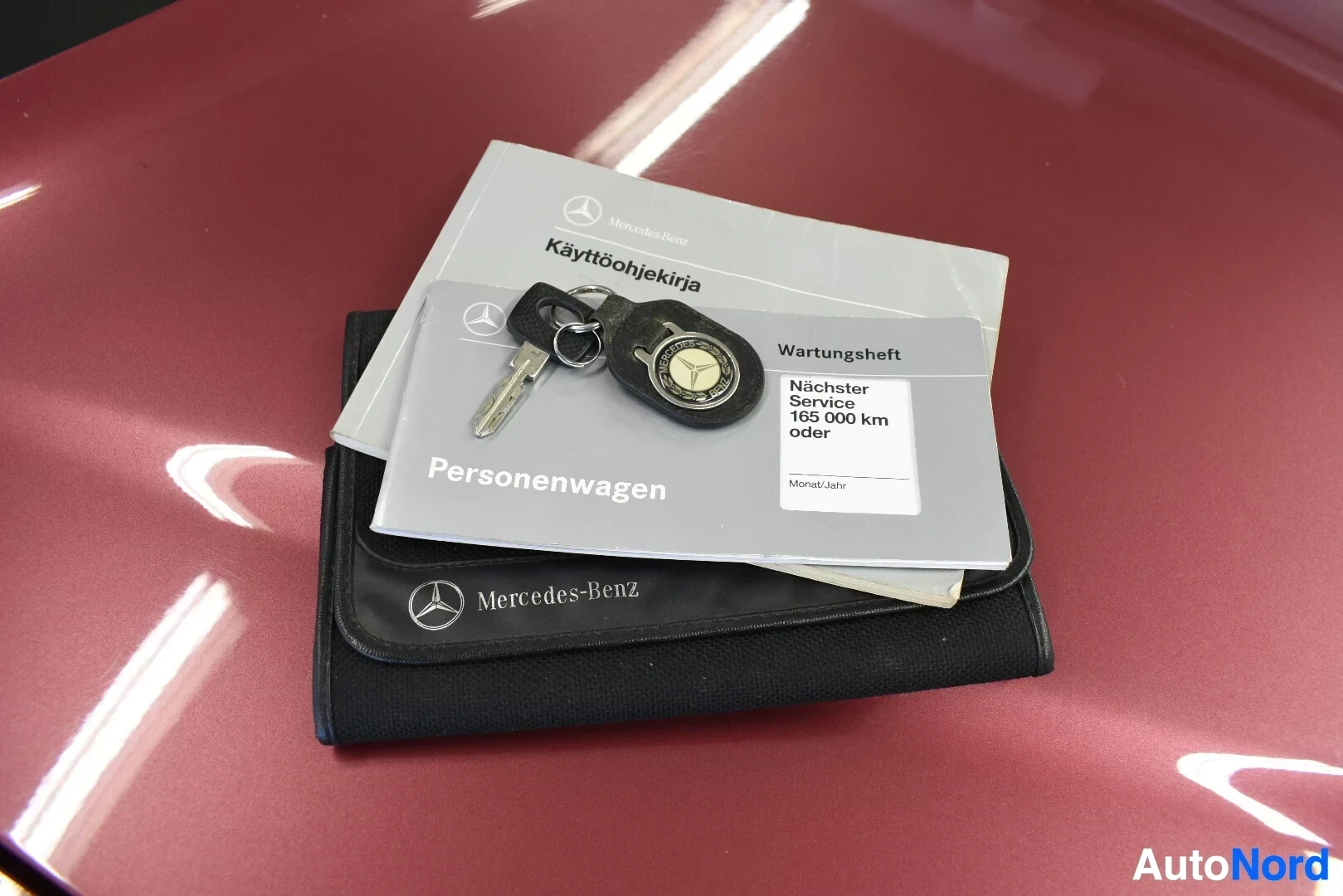 Asiakirja: Kuvassa on useita dokumentteja, auton avain ja avaimenperä. Yhdessä dokumentissa lukee "Mercedes-Benz Käyttöohjekirja" ja toisessa "Wartungsheft Nächster Servic