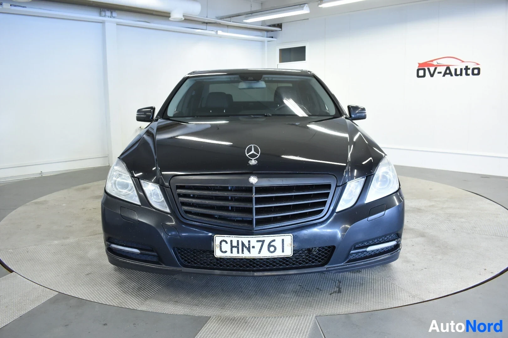 Edestä: Musta sedan-tyyppinen henkilöauto on kuvattu suoraan edestä. Auton etusäleikössä on Mercedes-Benz-logo ja etupuskurissa on rekisterikilpi ""