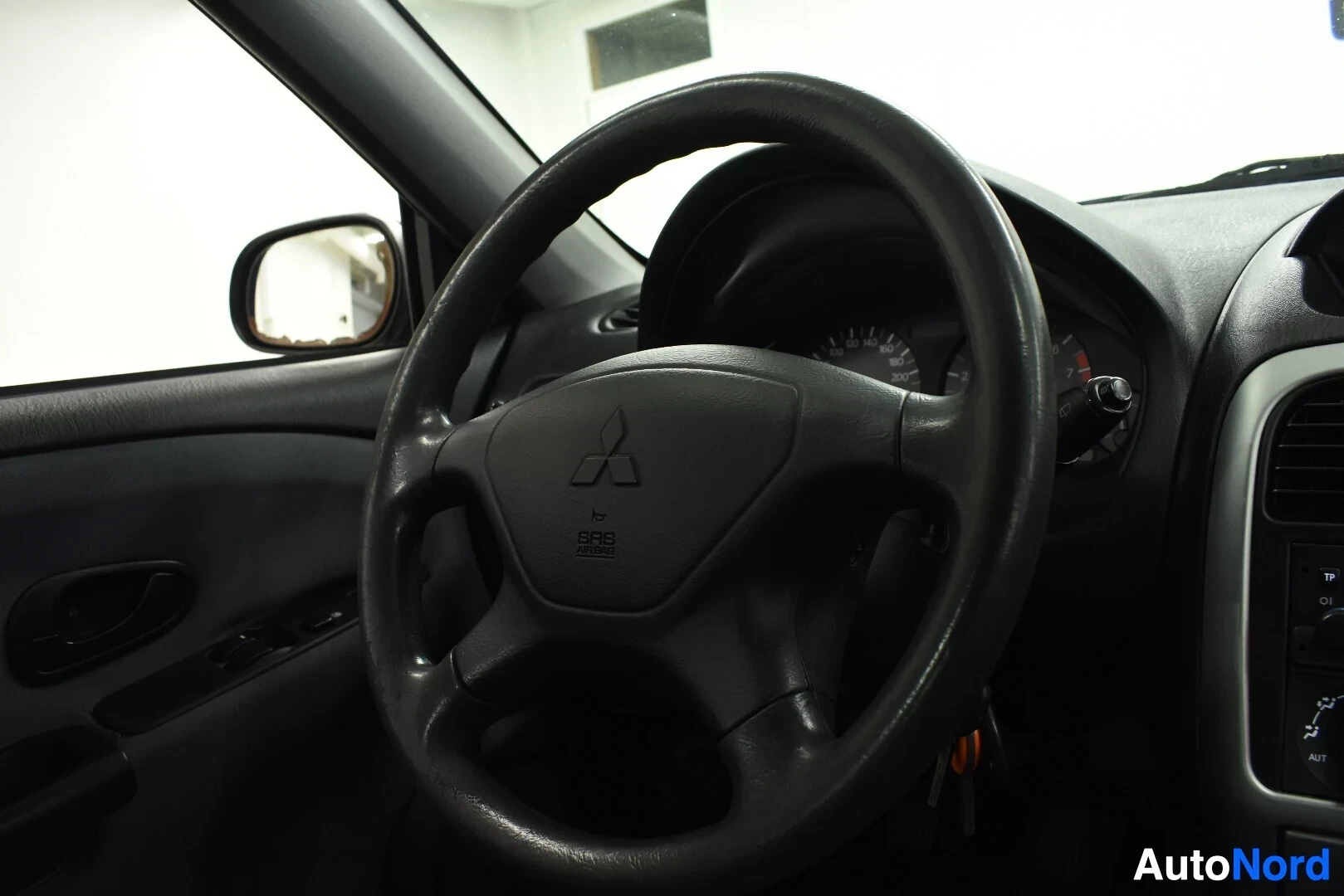Ohjaamo: Kojelaudan yksityiskohta ohjauspyörästä, jossa on Mitsubishi-logo ja SRS AIRBAG -merkintä, sekä mittaristoa.
