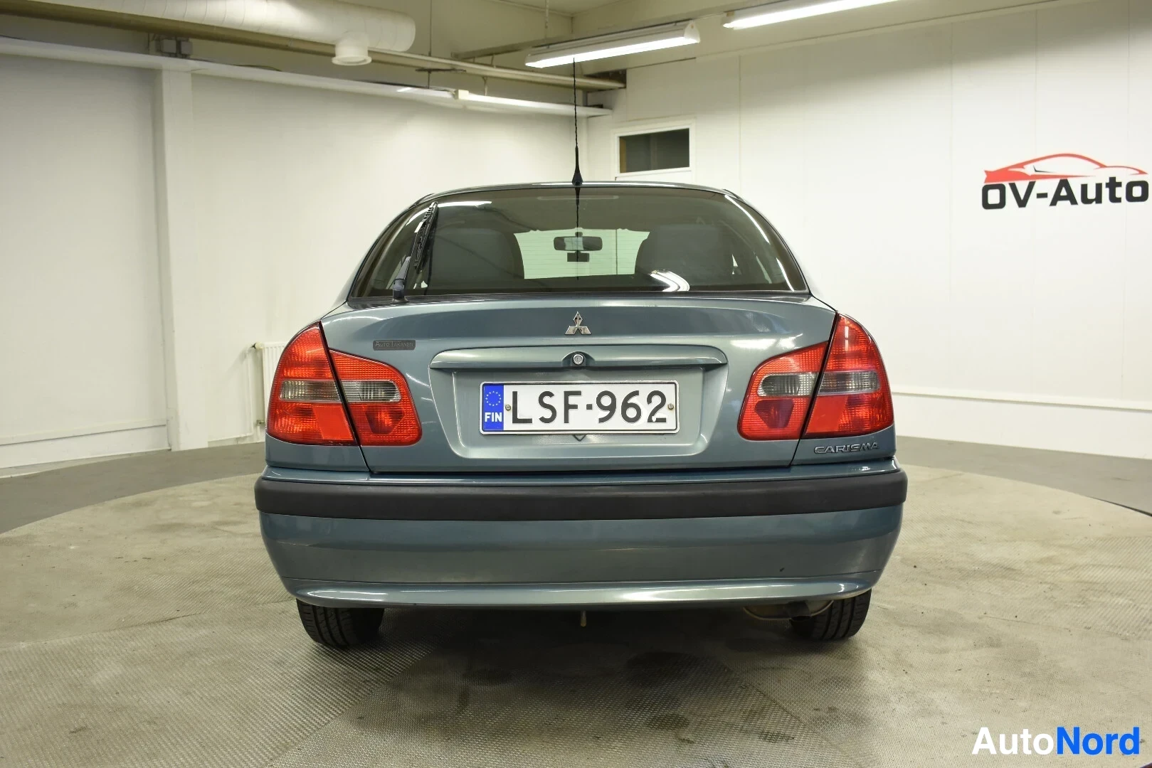 Takaa: Harmaa henkilöauto, jossa on tumma takapuskuri, on kuvattu suoraan takaa. Takavalot ja takarekisterikilpi ovat näkyvissä. Oikealla on sinivalkoinen logo OV-Auto